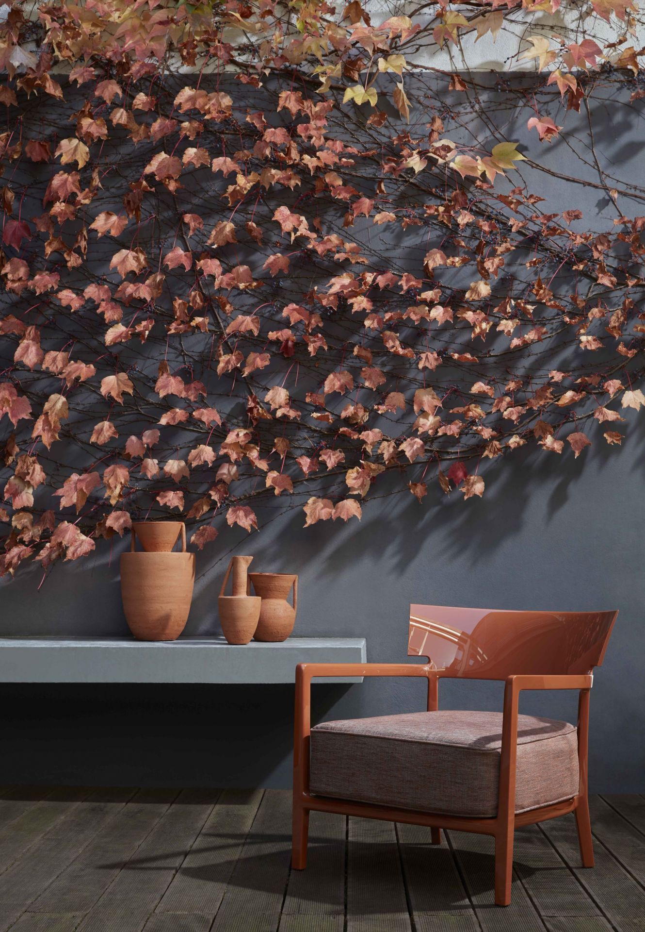 Cara Outdoor Sessel von Kartell in Terrakotta vor grauer Wand mit Efeu und Vasen.