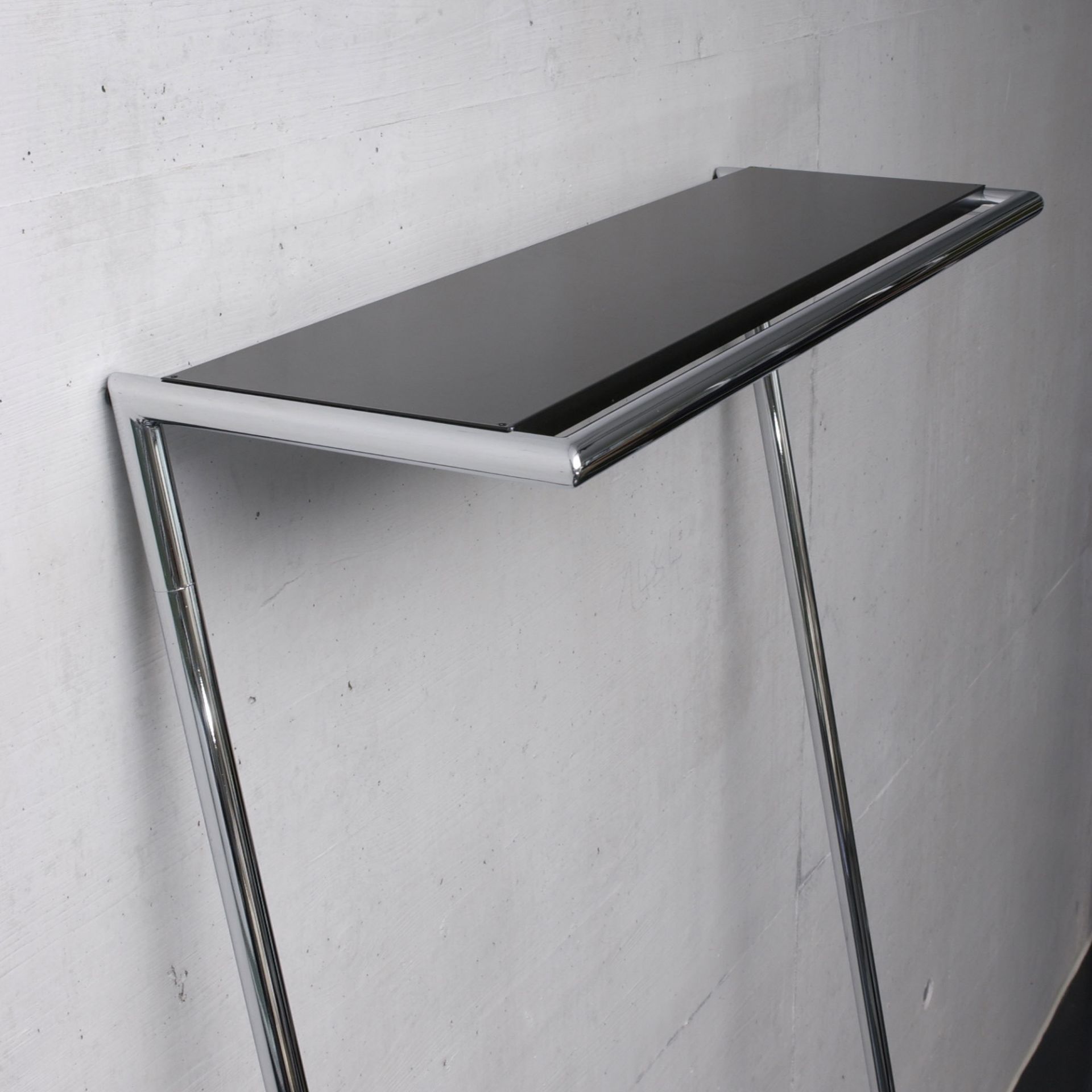 Schwarze Hutablage für Zen und Link Garderobe von Mox mit verchromtem Gestell vor einer Betonwand.