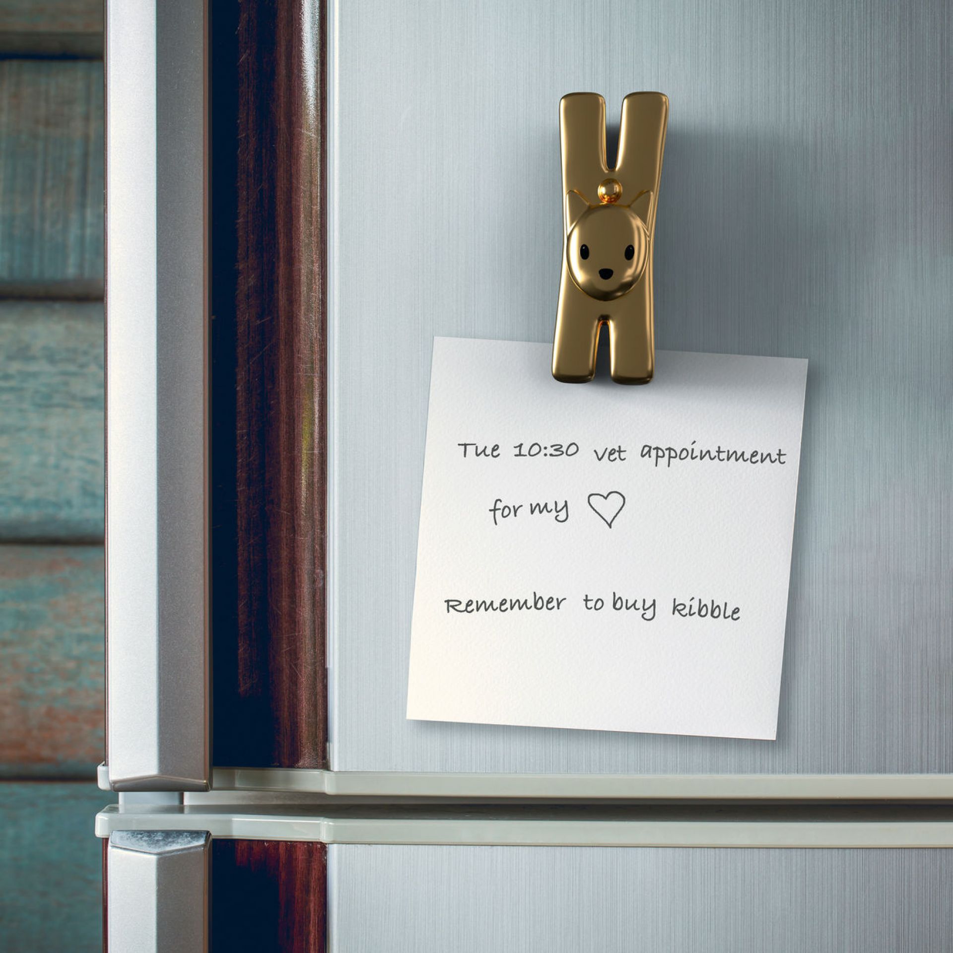 Goldfarbene Klammer mit Tiergesicht hält Notiz auf Kühlschrank. Notizzettel erinnert an Tierarzttermin und Tierfutter.