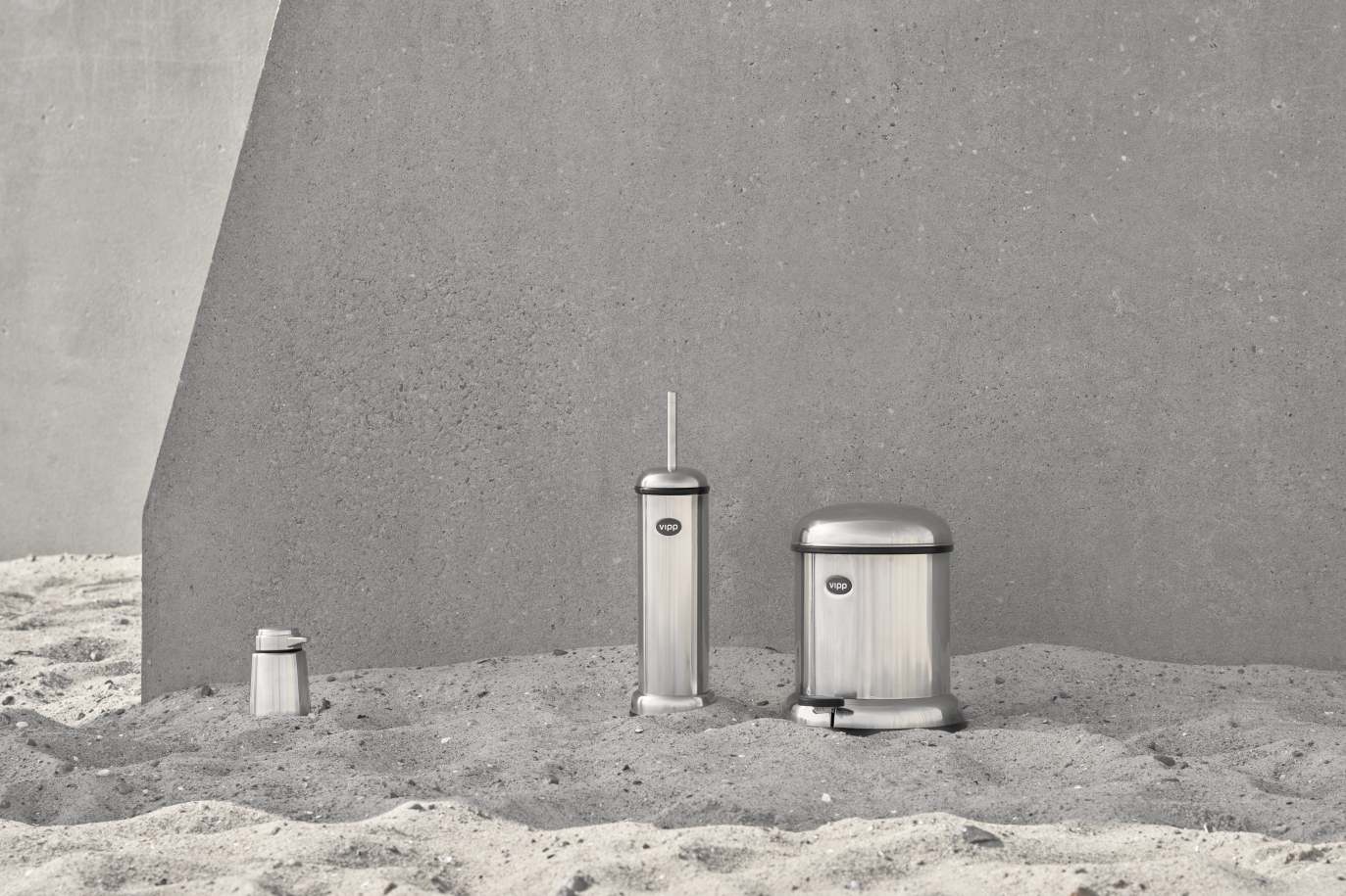 Vipp Badezimmer-Set aus Edelstahl bestehend aus Seifenspender, Toilettenbürste und Treteimer vor grauer Wand.