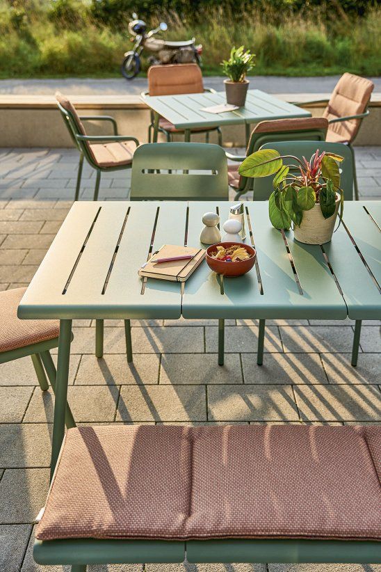 Weishäupl Vapio Stool Hocker Outdoor: Gartentisch mit Stühlen und Bank in Grün, gedeckt mit Schüssel, Notizbuch und Pflanze.