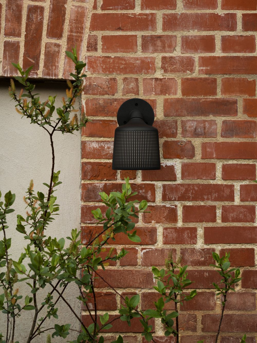 Schwarzer Vipp Wandspot für Außenmontage an einer roten Backsteinmauer, umgeben von Grünpflanzen.