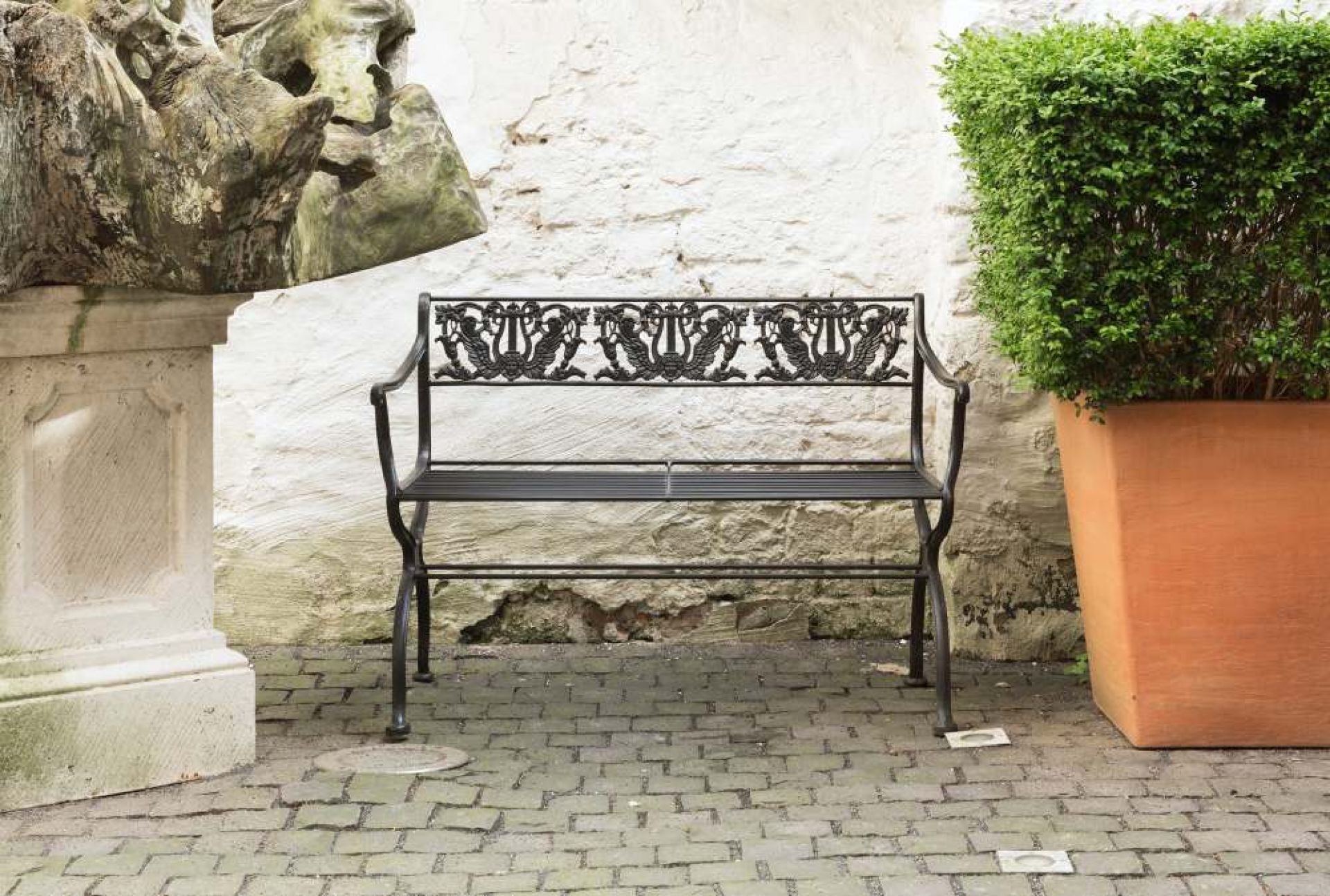 Schwarze Tecta Gartenbank mit filigranem Muster vor weißer Wand und Pflanzkübel.