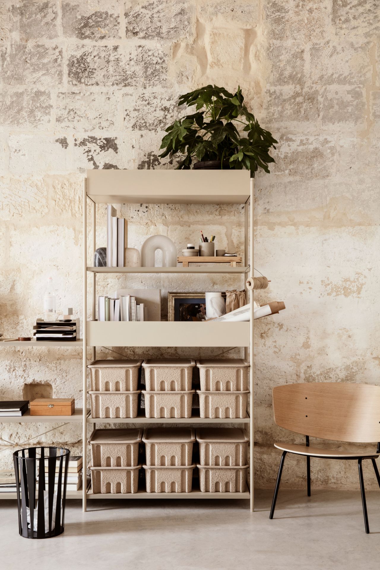 Beige Ferm Living Regalsystem mit Aufbewahrungsboxen, Dekoration und Pflanze vor einer strukturierten Wand.
