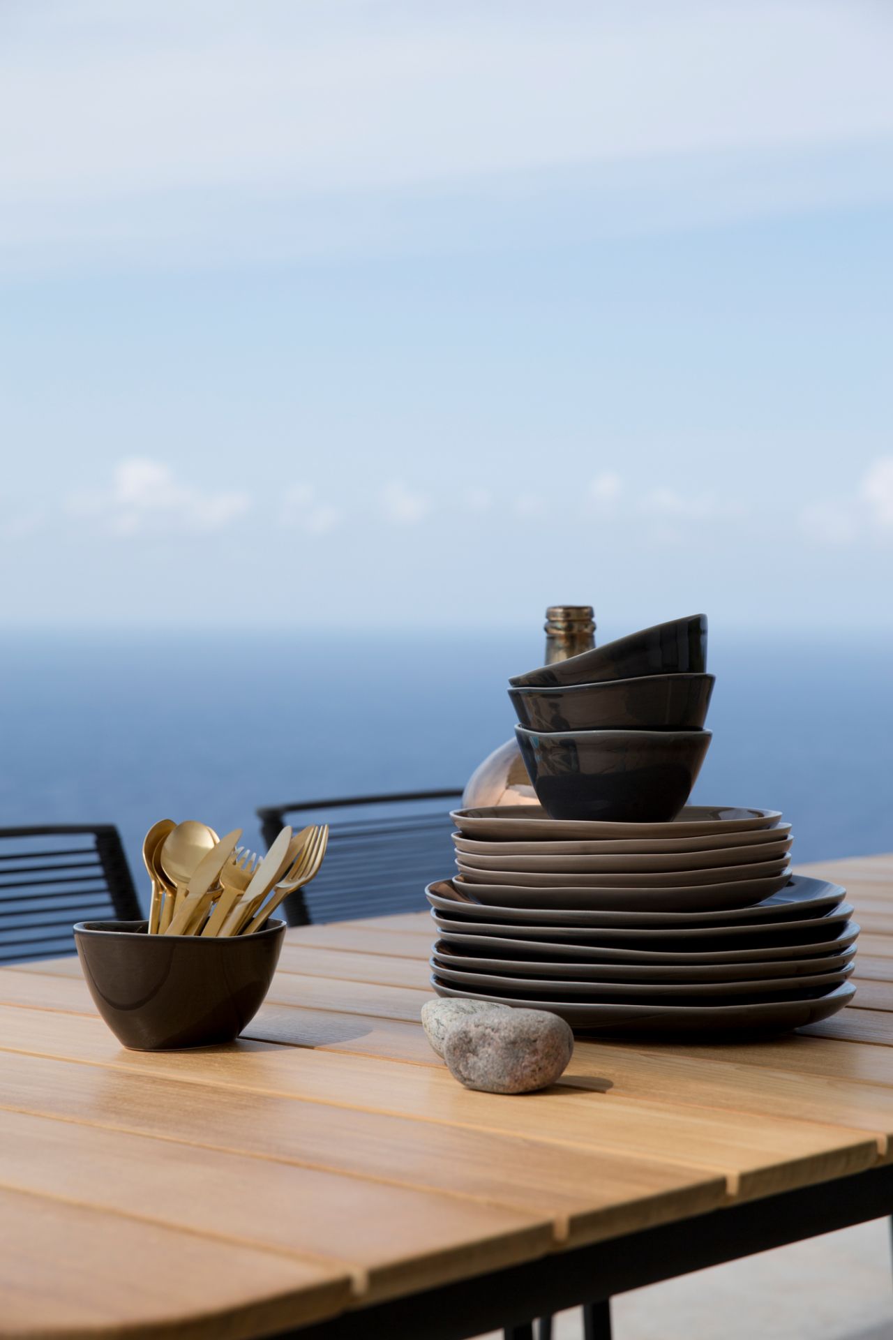 Cane-Line Core Outdoor Esstisch Teak mit Geschirr und goldenem Besteck vor Meereskulisse.