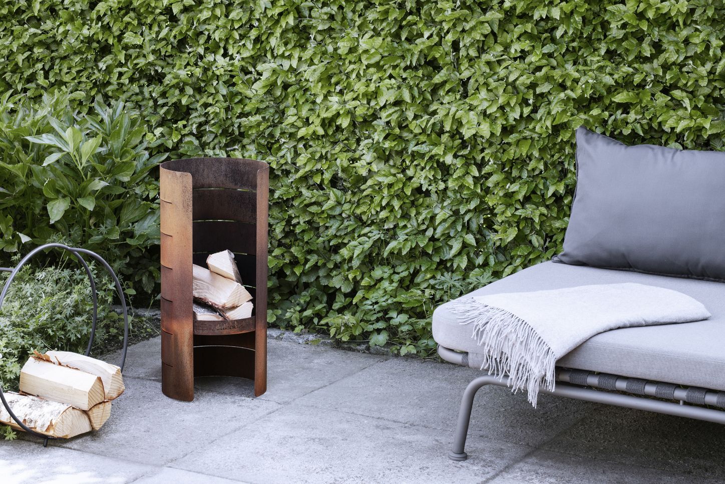 Eva Solo FireCylinder Gartenkamin aus Roststahl mit Holzscheiten und Lounge-Möbeln im Garten.