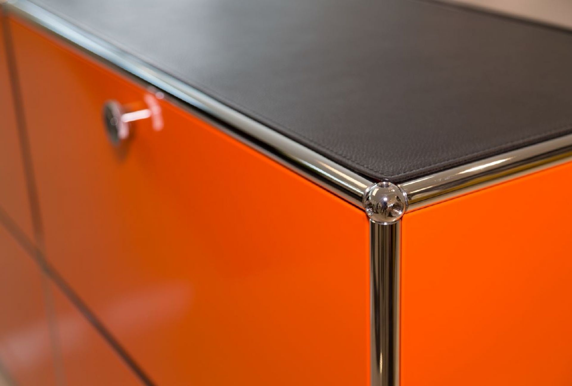 Detailaufnahme: Graphitschwarze Lederauflage auf orangefarbenem USM Sideboard mit Chrom-Gestell. Hochwertige Ledermatte für Möbel.