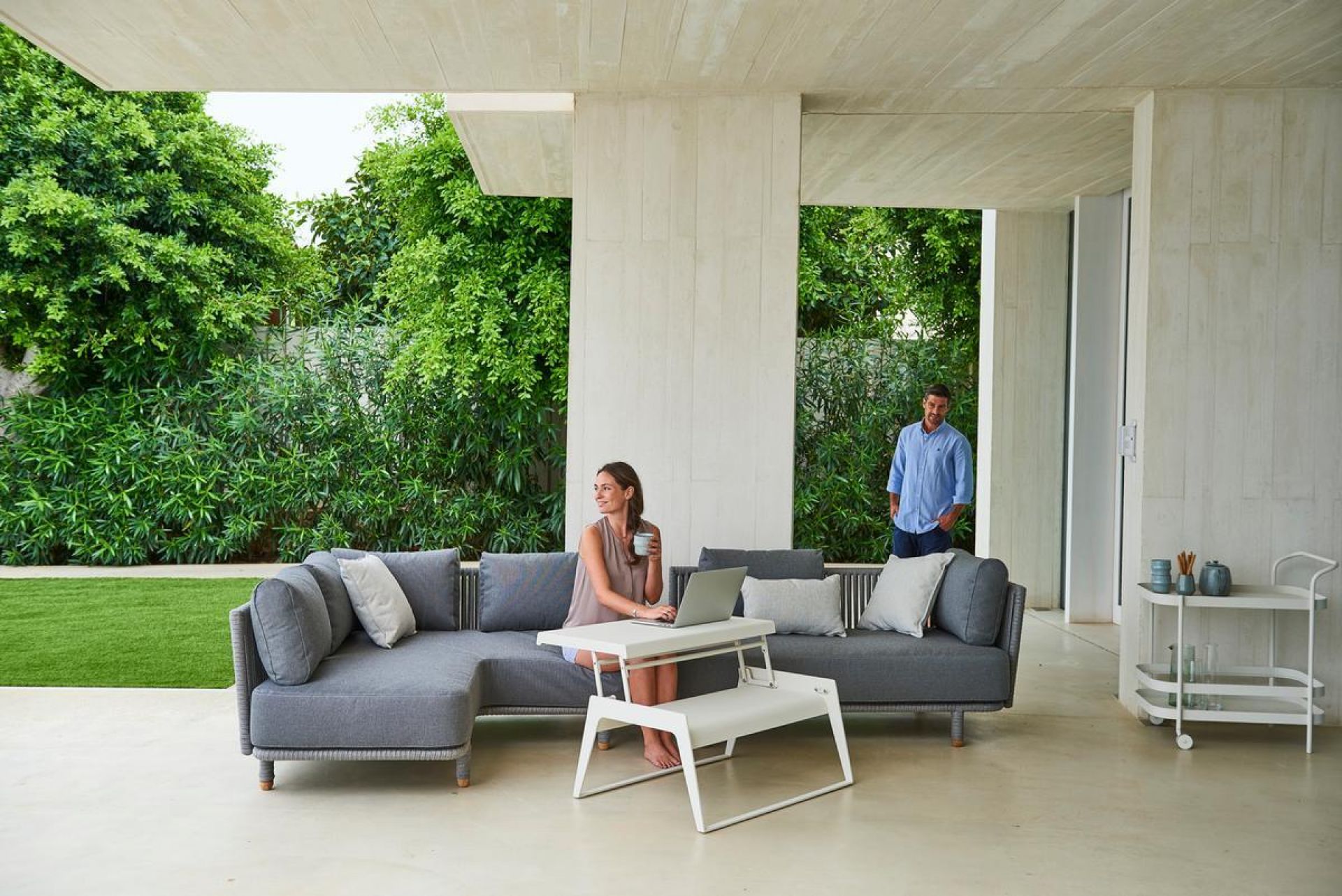 Frau nutzt Laptop auf Cane-Line Moments Outdoor Sofa. Graues 2-Sitzer Gartensofa mit Kissen.