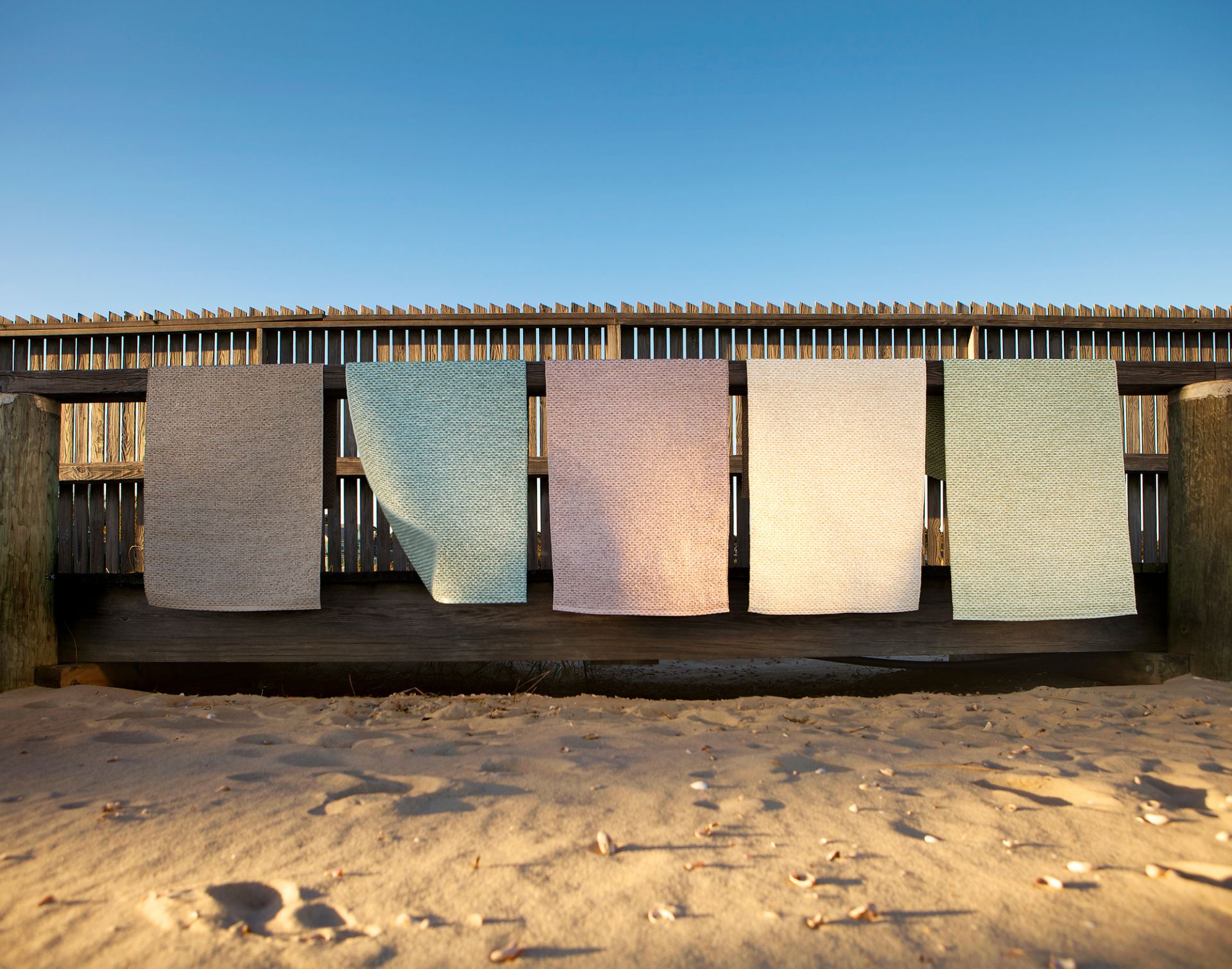 Svea Kunststoffteppiche von Pappelina, 70x160 cm, in verschiedenen Farben hängen auf einer Holzbank am Strand.