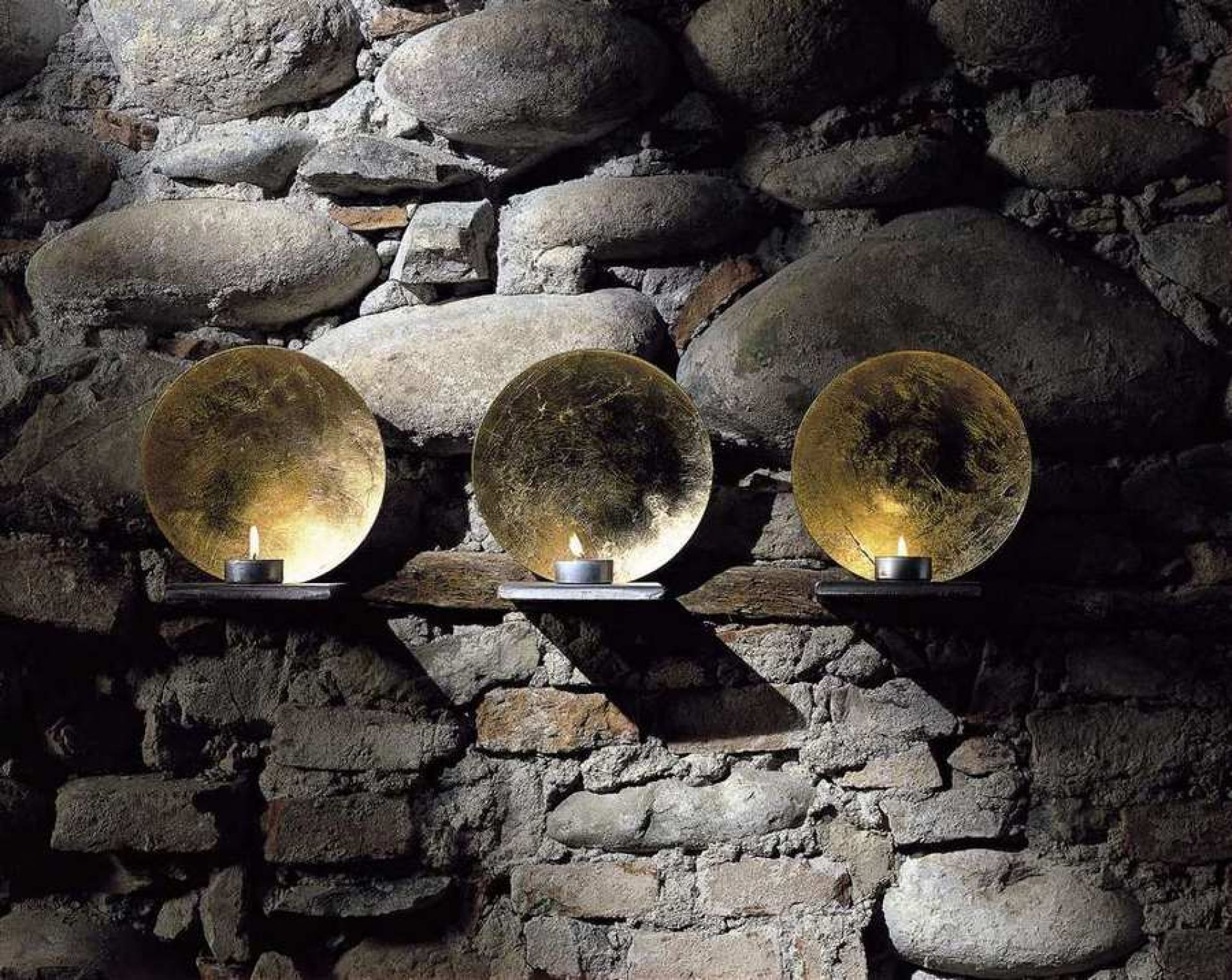 Drei Luna Kerzenhalter von Catellani & Smith mit Teelichtern vor einer Steinwand. Goldene Lichtobjekte.