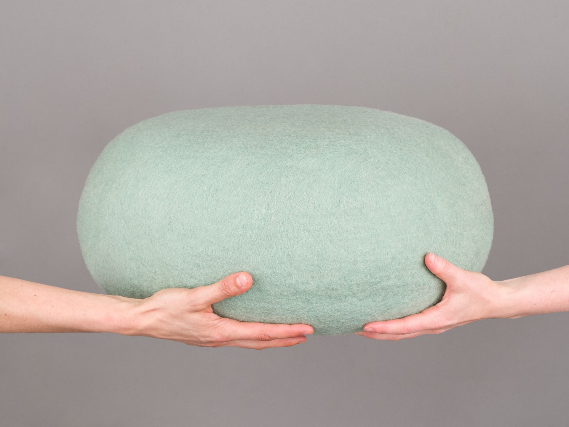 Hände halten einen runden, hellgrünen Filz Pouf Hocker. Gemütliches Wohnaccessoire für Sitzgelegenheiten.