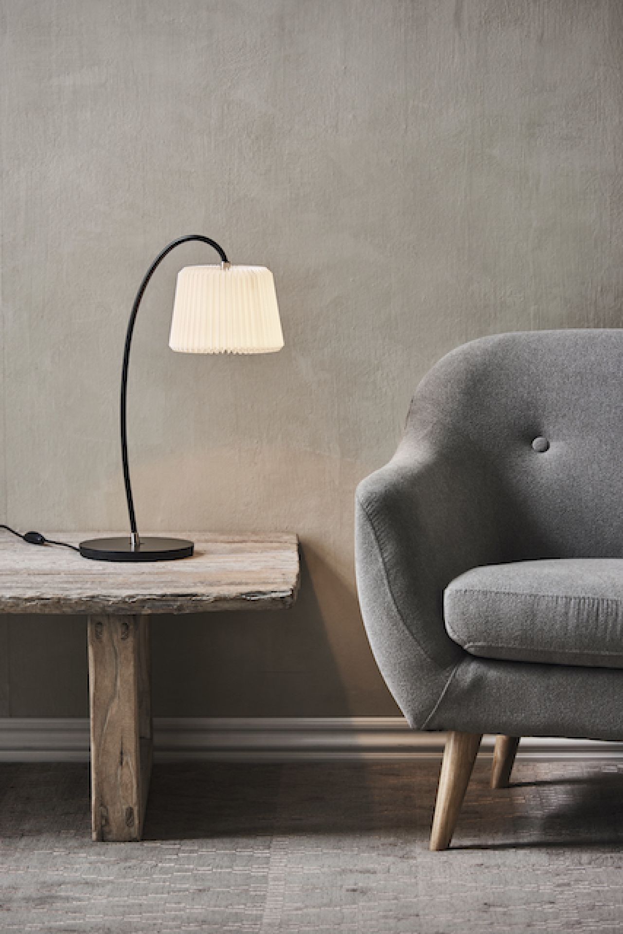 Snowdrop Tischleuchte von Le Klint mit plissiertem Schirm auf einem Holztisch neben einem Sessel.