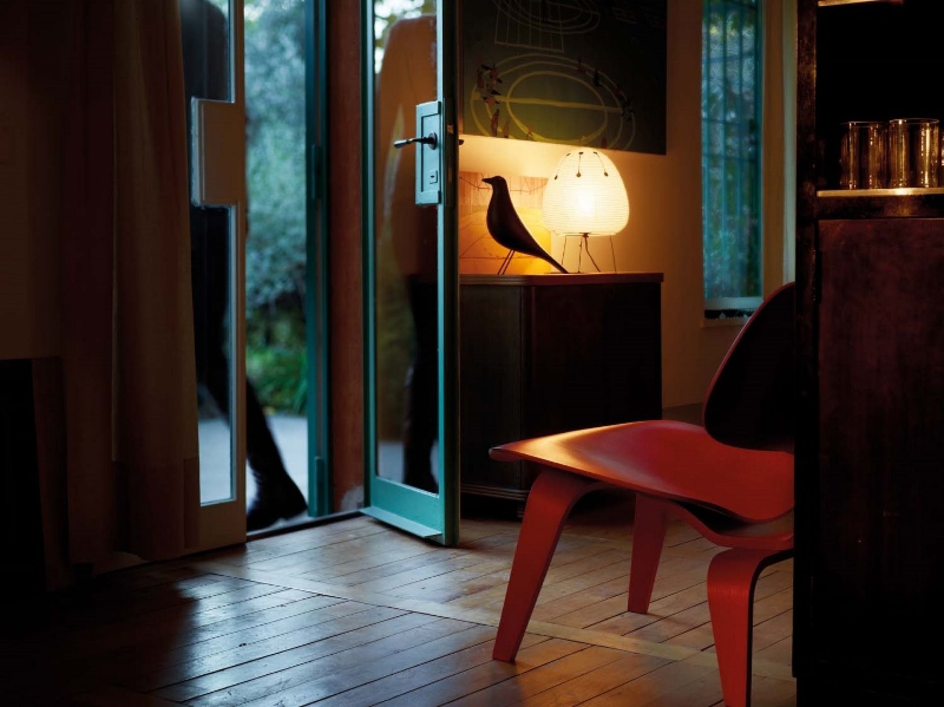 Interieur mit Eames House Bird Skulptur, Lampe und rotem Stuhl auf Holzboden. Dekorative Wohnaccessoires.