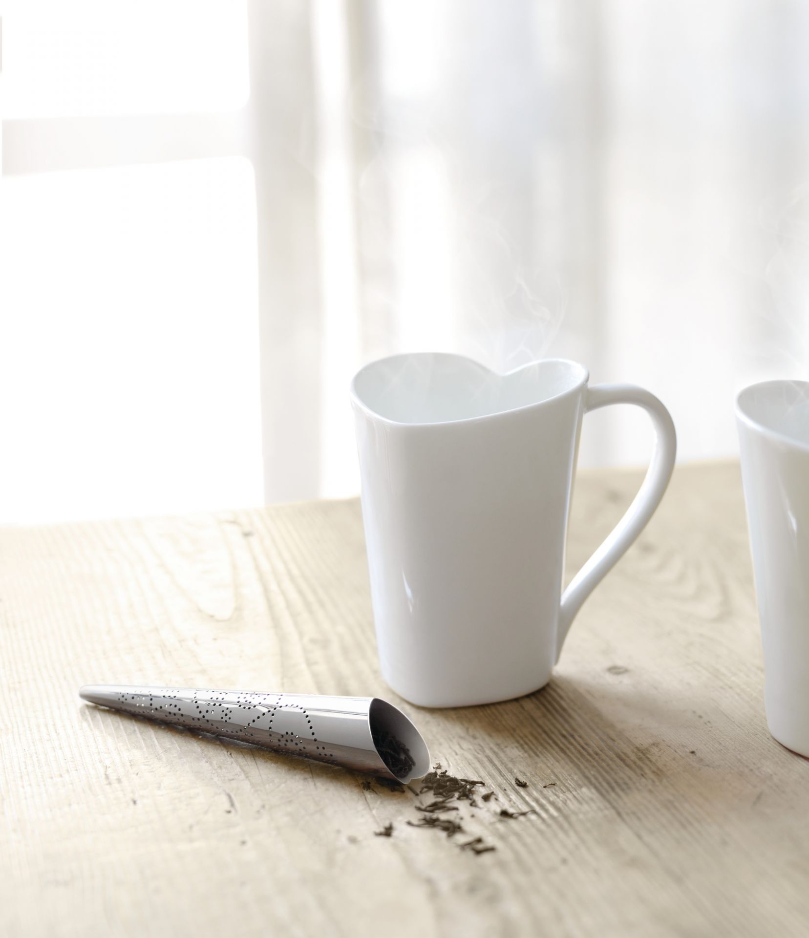 Produktfoto: Weiße Alessi Tasse mit Herzform und Teesieb auf hellem Holztisch.