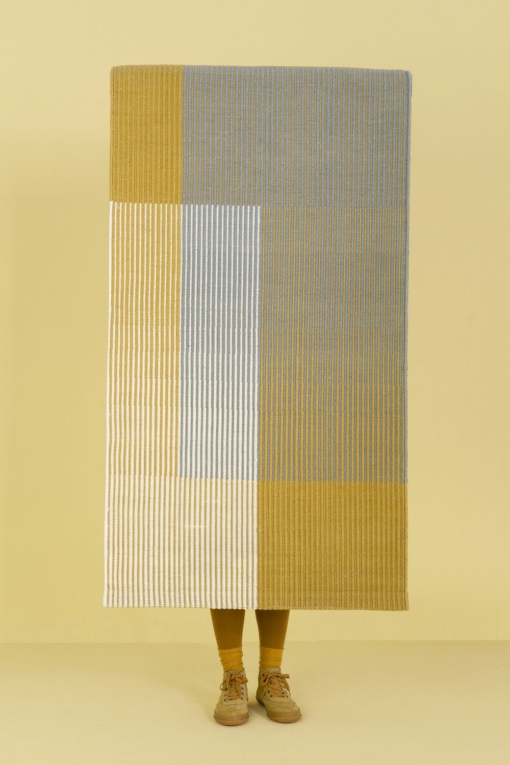 Nanimarquina Haze Rug Teppich mit geometrischem Muster in Gelb, Grau und Weiß, stehend vor Person.