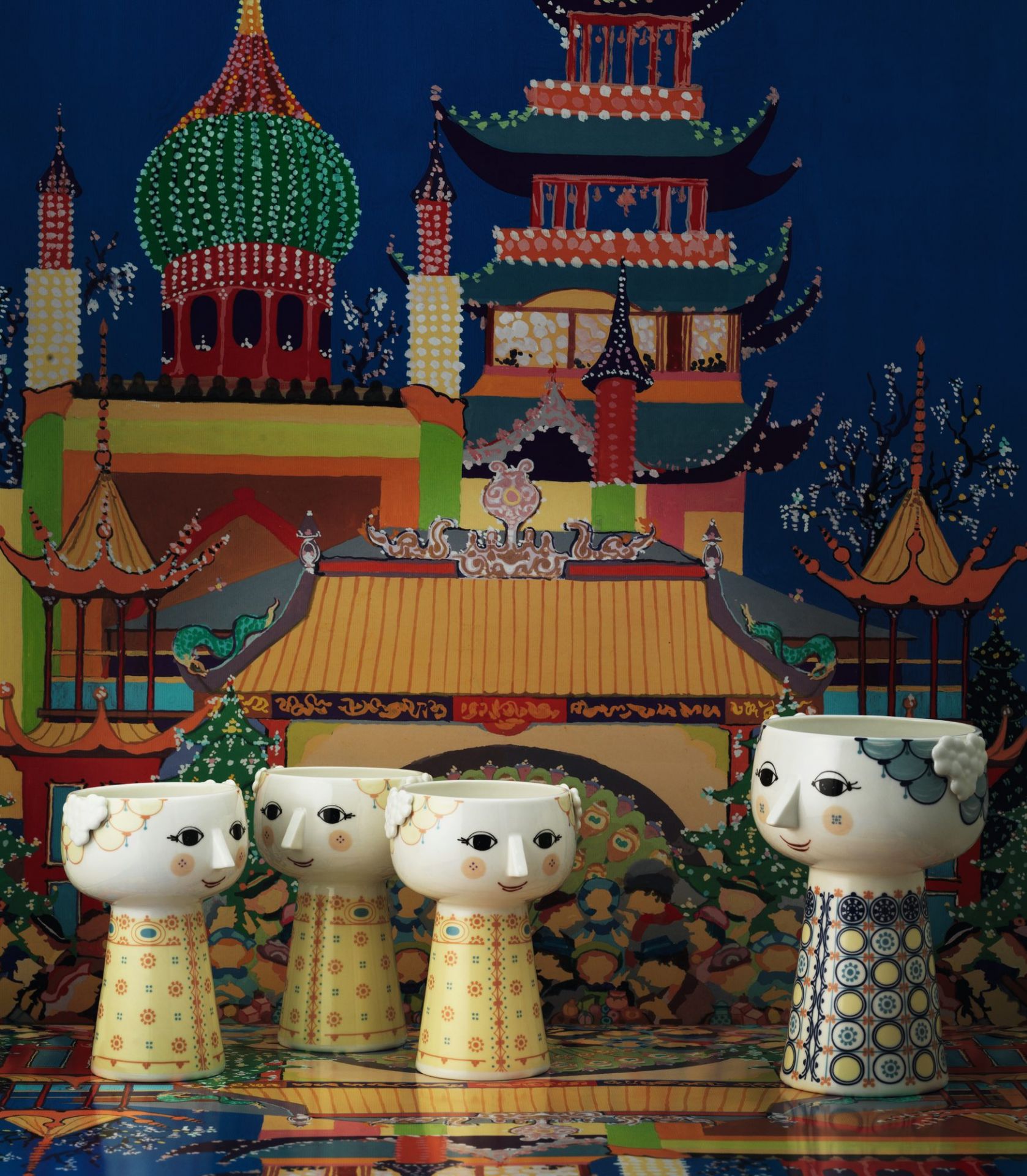 Gelbe Bjorn Wiinblad Eva Vase, 15 cm, mit Gesicht und floralem Muster vor farbenfrohem Hintergrund.