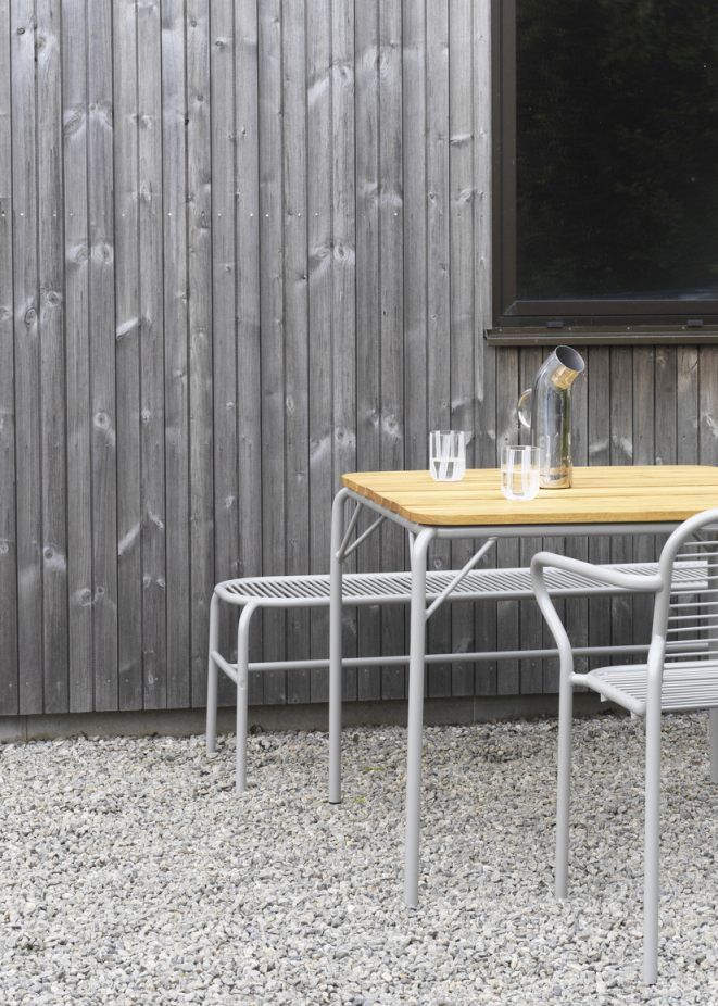 Normann Copenhagen Vig Bench Bank Outdoor mit Tisch und Gläsern vor grauer Holzwand.