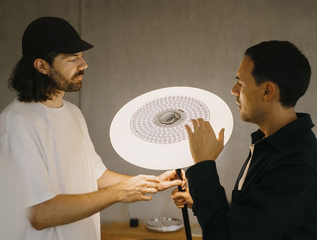 Zwei Personen betrachten eine Oto Stehleuchte mit Rundfuß und warmweißem Licht.