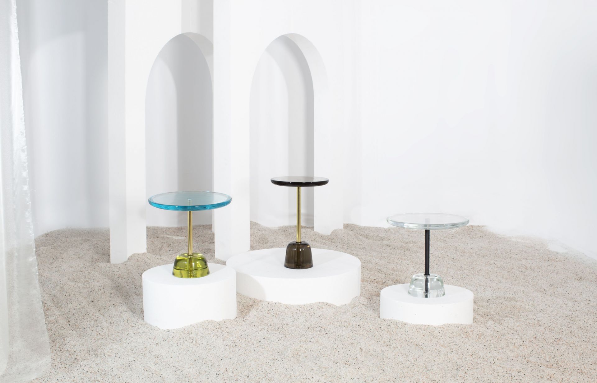 Drei moderne Beistelltische von Pulpo mit Glasplatte und farbigen Sockeln auf Sand.