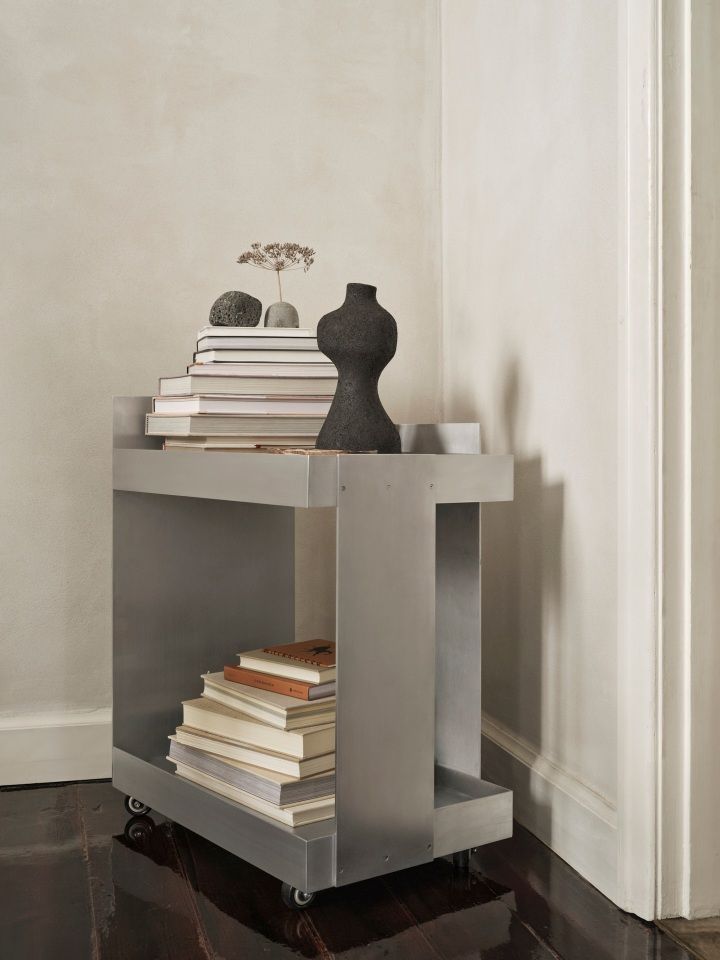 Ferm Living Lager Trolley: Aluminium Servierwagen mit zwei Ablagen, dekoriert mit Büchern und Deko-Objekten.