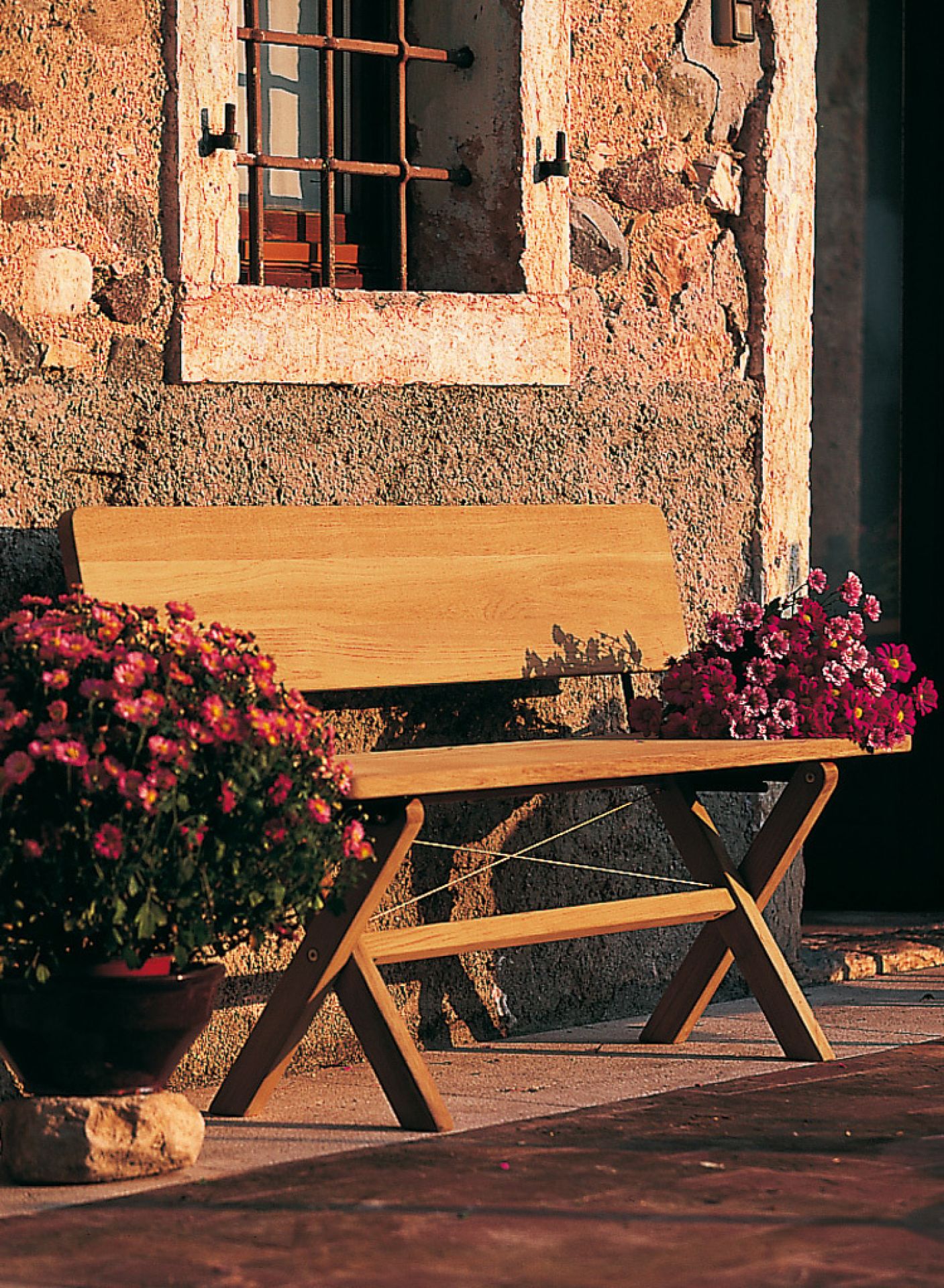 Weishäupl Cross Bank klappbar aus Holz mit Rückenlehne und Blumen vor Steinmauer.