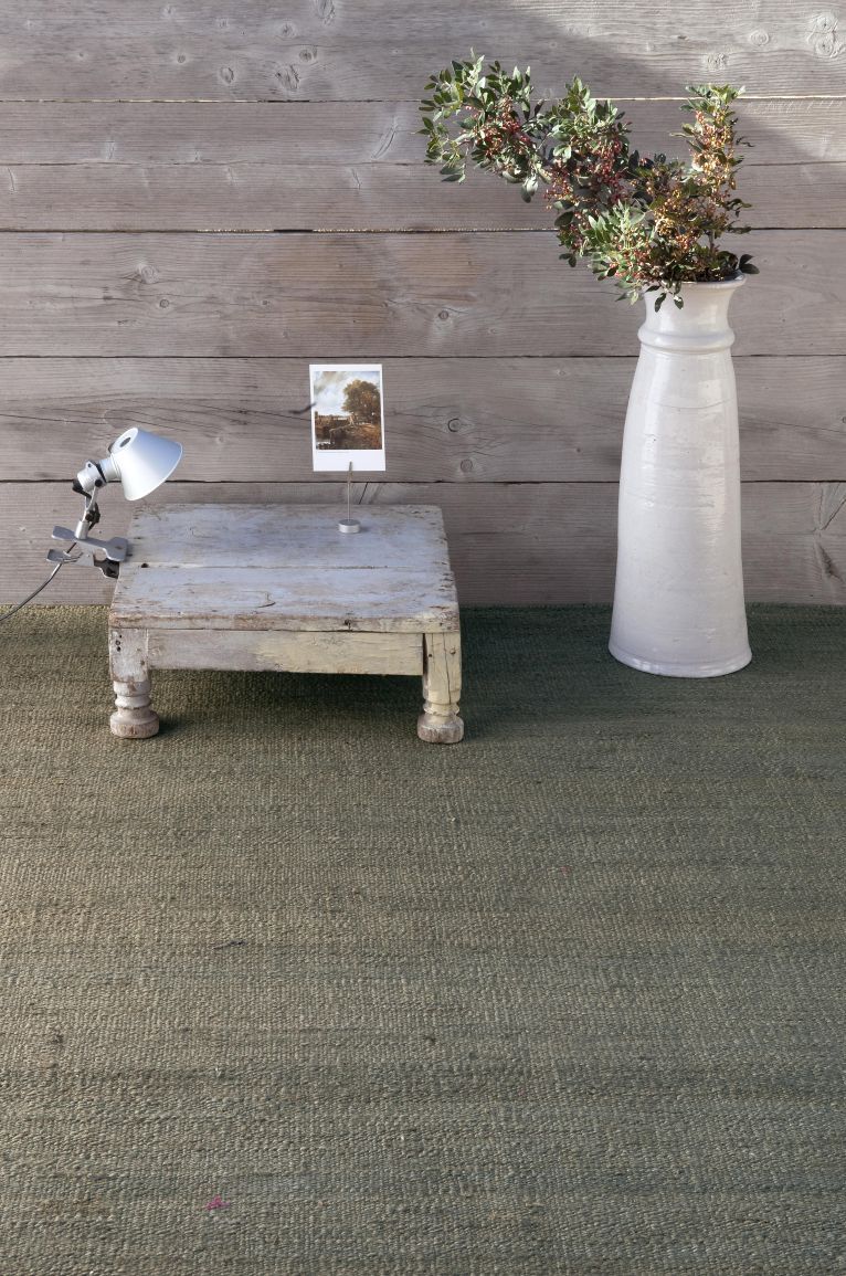 Nanimarquina Vegetal Teppich Natural 200x300cm, Detailaufnahme des grünen Naturfaser Teppichs mit Holzdekor und Vase.