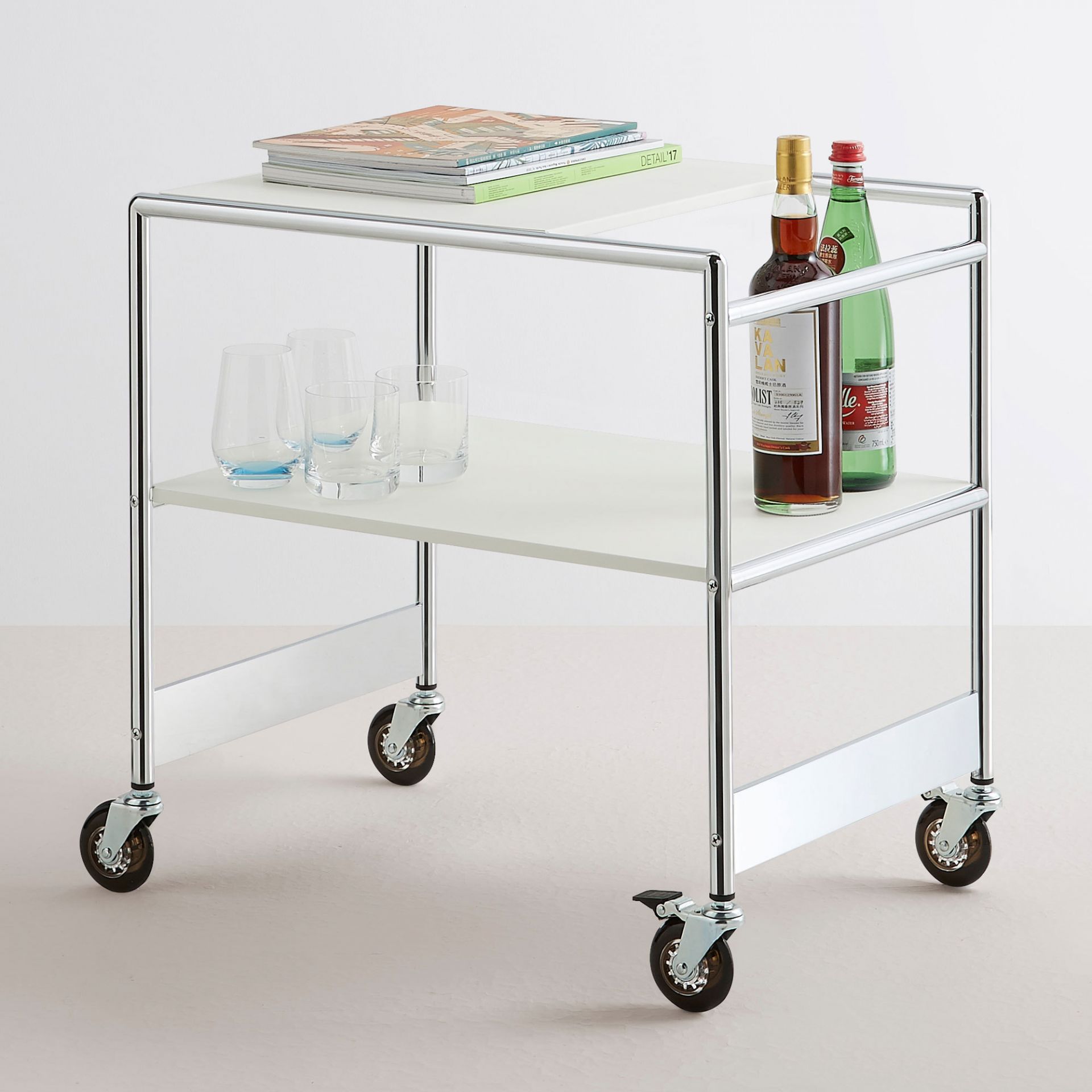 SIR Servierwagen mit zwei weißen Ablageflächen und Chromgestell, mit Gläsern, Getränken und Zeitschriften.