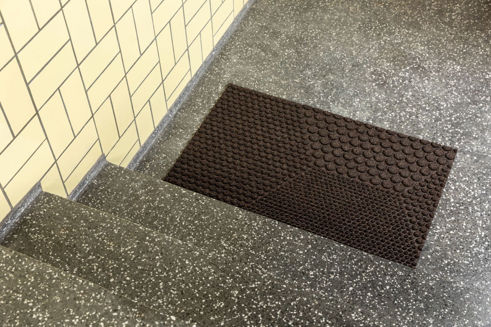 Braune Hex Fußmatte Soil von Heymat auf einer Treppe mit Terrazzo-Boden und gelben Fliesen.
