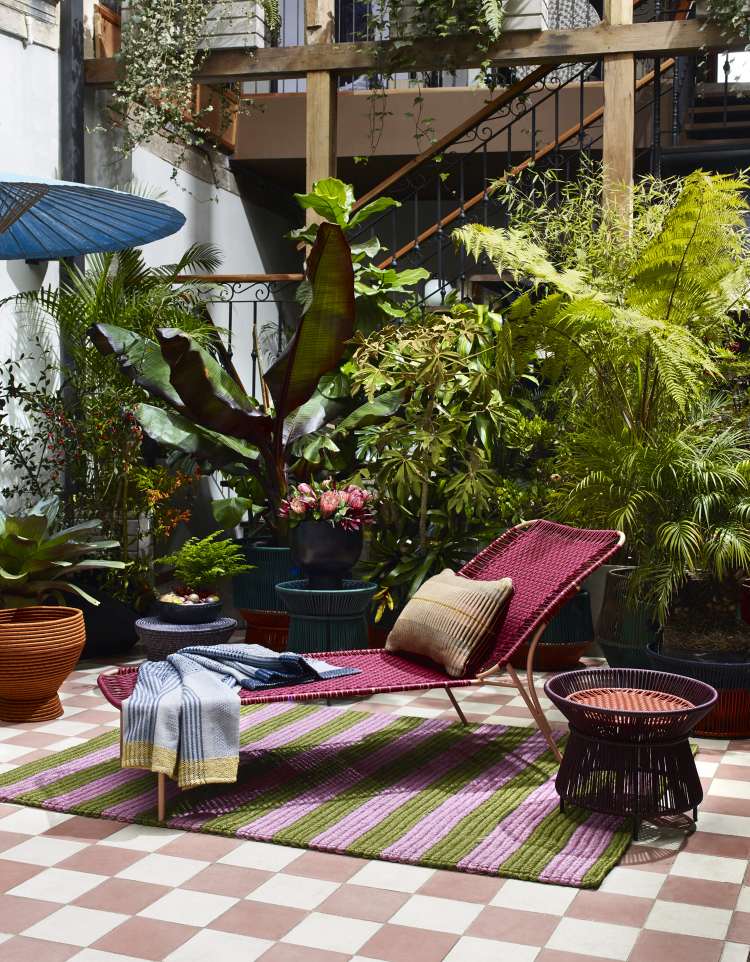 Ames Caribe Chic Basket Table als Outdoor-Beistelltisch neben einer Liege im Garten.