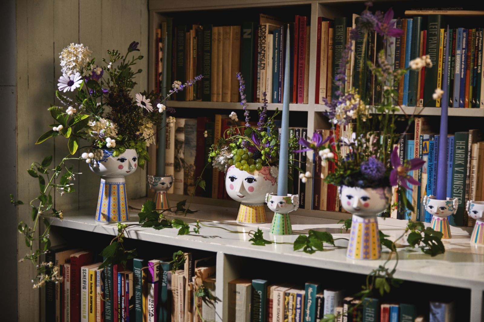 Keramikvase Eva H15cm Lavendel von Bjørn Wiinblad mit Blumen und Kerzen auf einem Bücherregal.