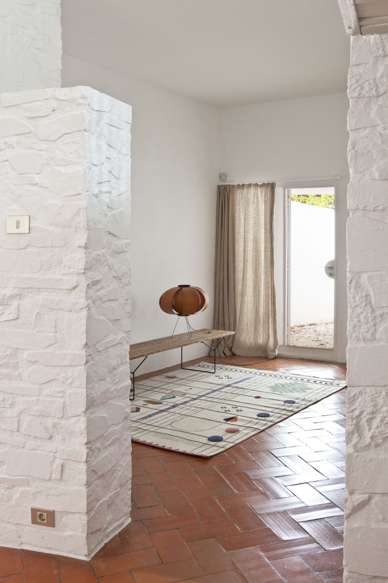 Rabari Teppich von Nanimarquina in hellem Raum mit Terrakotta-Fliesen und weißer Backsteinwand.