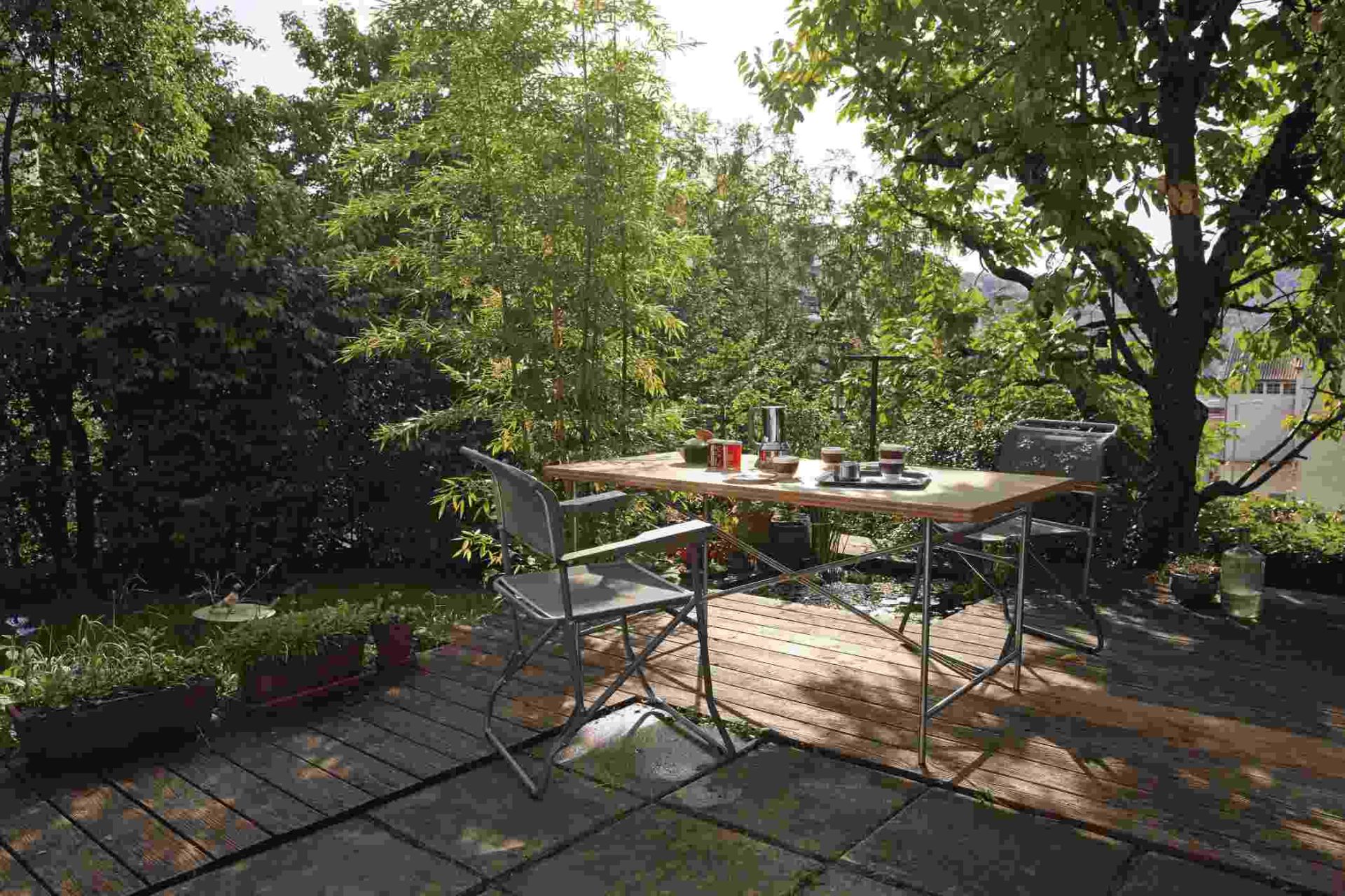 Eiermann Tisch mit zwei Stühlen auf einer Terrasse mit Pflanzen und Bäumen im Hintergrund.