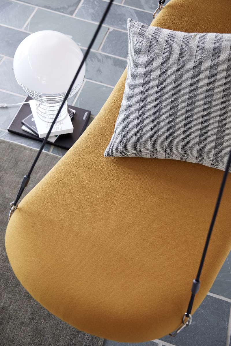 Detailaufnahme eines gelben Hängesessels mit gestreiftem Kissen, Lampe und Büchern im Hintergrund.