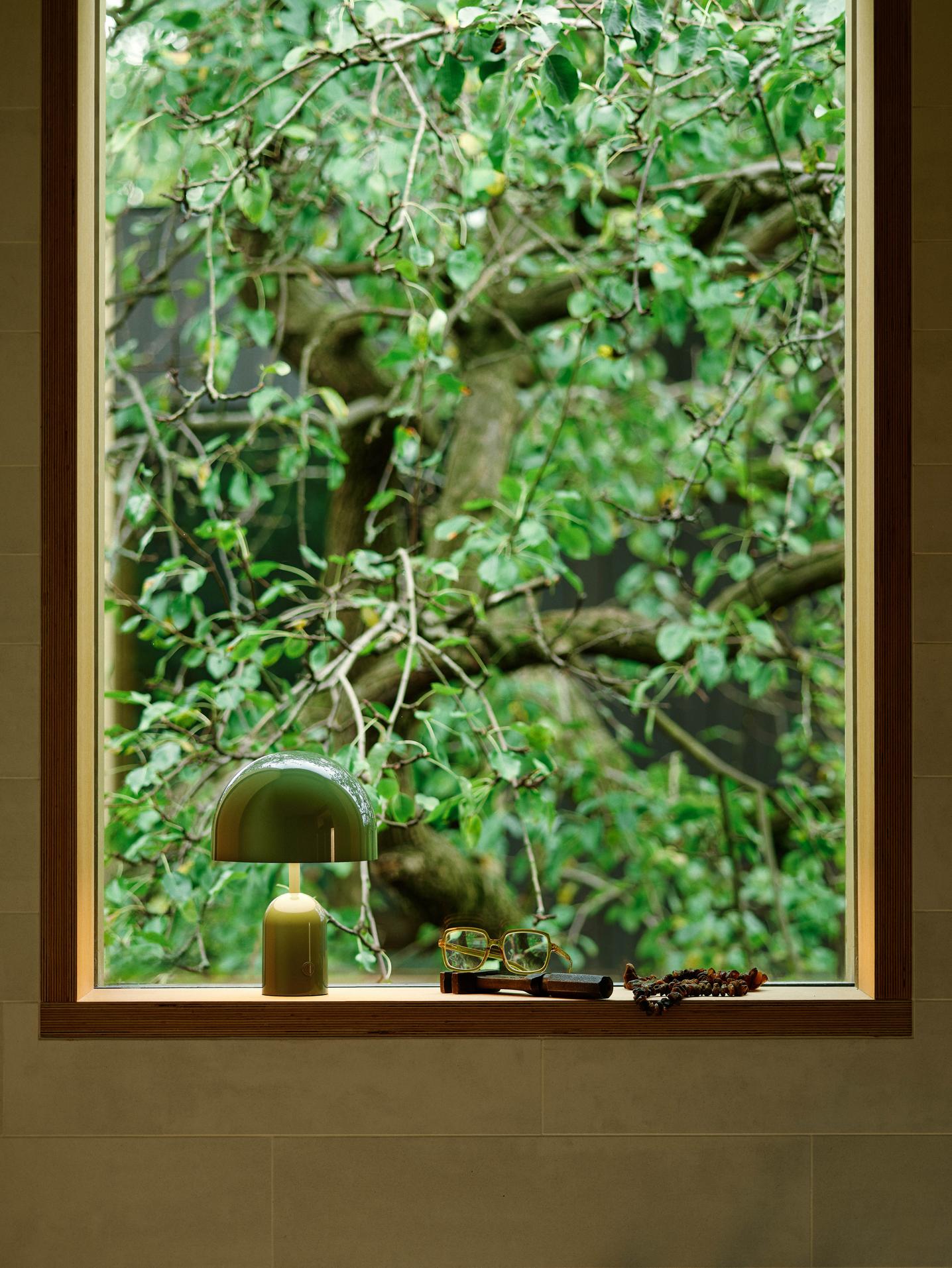 Grüne Bell LED Akkuleuchte von Tom Dixon auf Fensterbank mit Baum im Hintergrund.