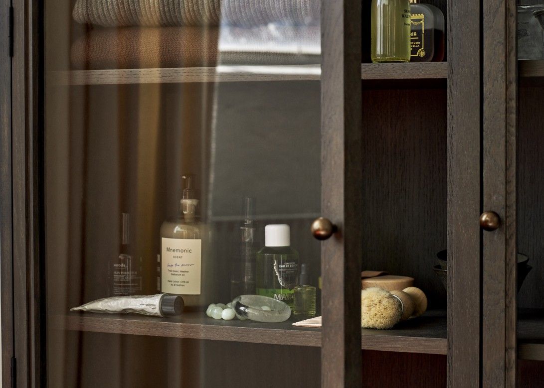 Detailaufnahme des AndTradition Trace SC88 Schranks mit Glasfront, gefüllt mit Kosmetikprodukten und Textilien.