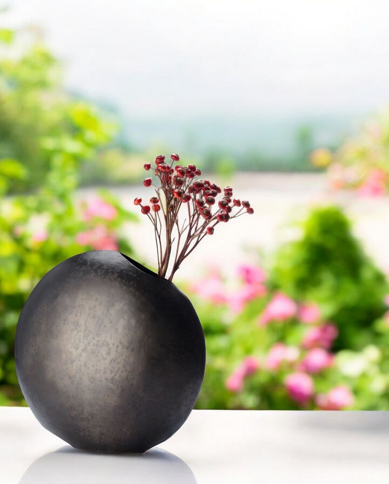 Monrovia Vase Philippi: Runde, dunkle Design-Vase mit roten Beeren-Zweigen vor verschwommenem Gartenhintergrund.