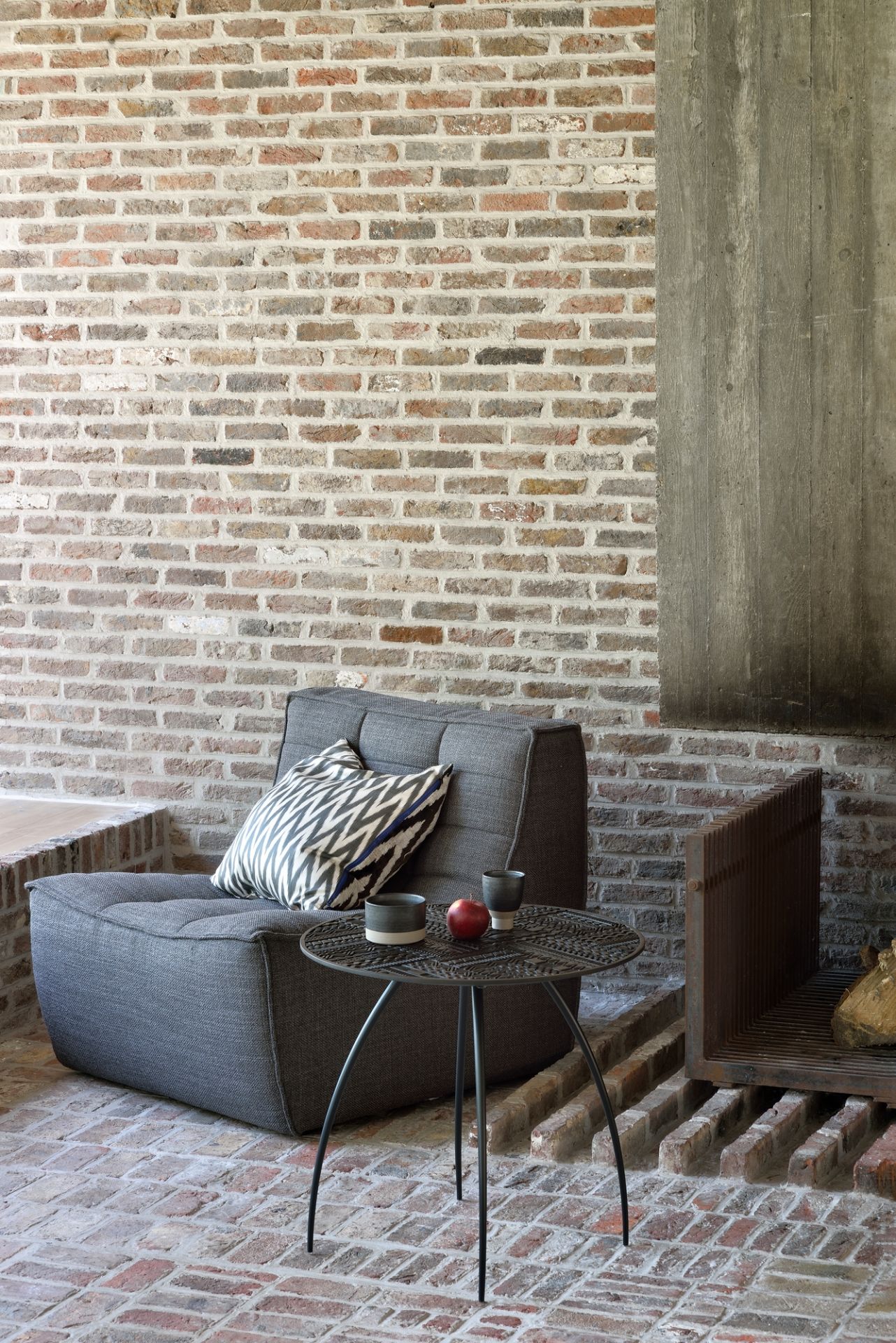 Grauer Sessel von Ethnicraft mit Kissen und Beistelltisch vor einer Backsteinwand.