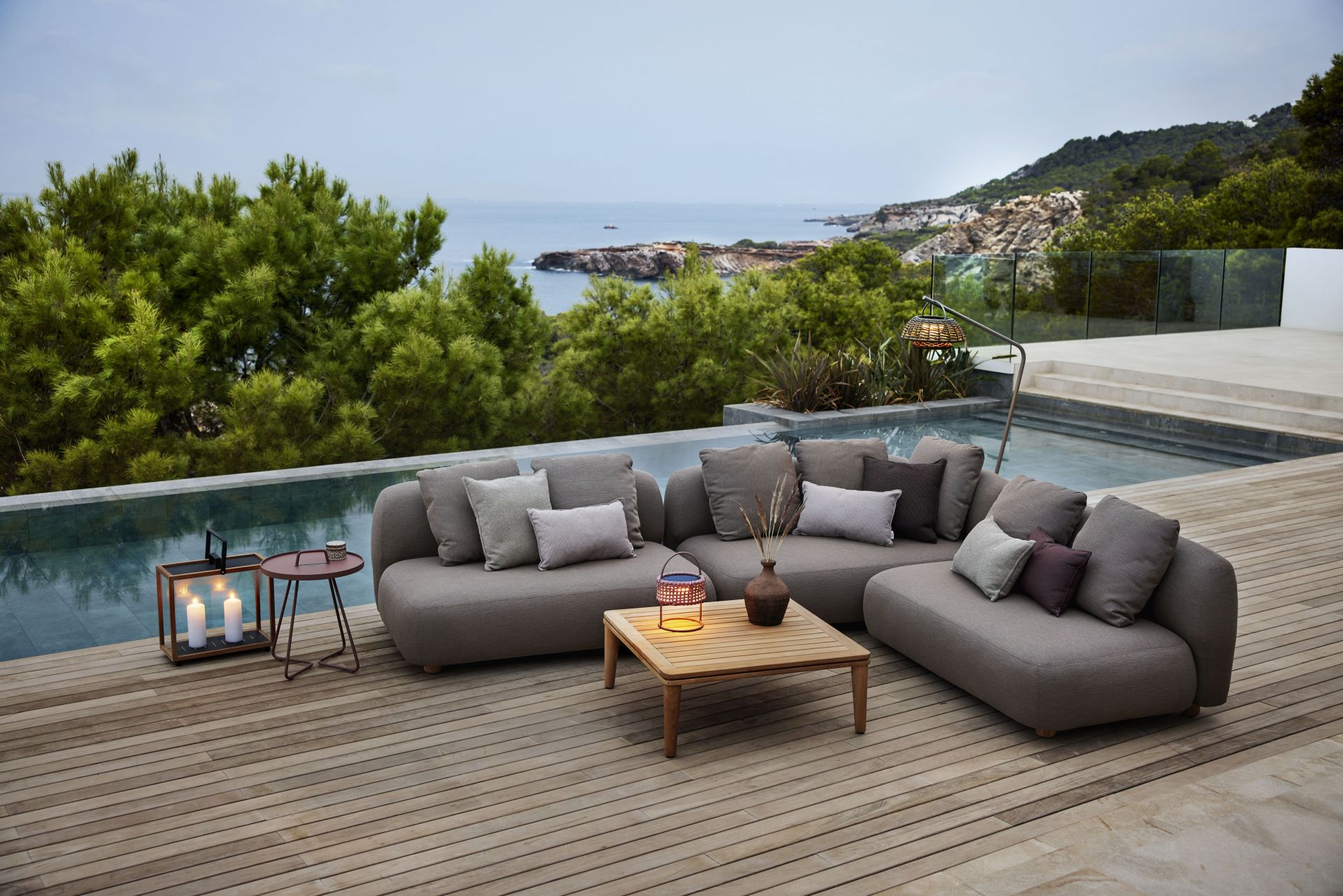 Cane-Line Capture Outdoor Couchtisch aus Teakholz neben grauer Lounge-Garnitur mit Kissen auf Holzterrasse.