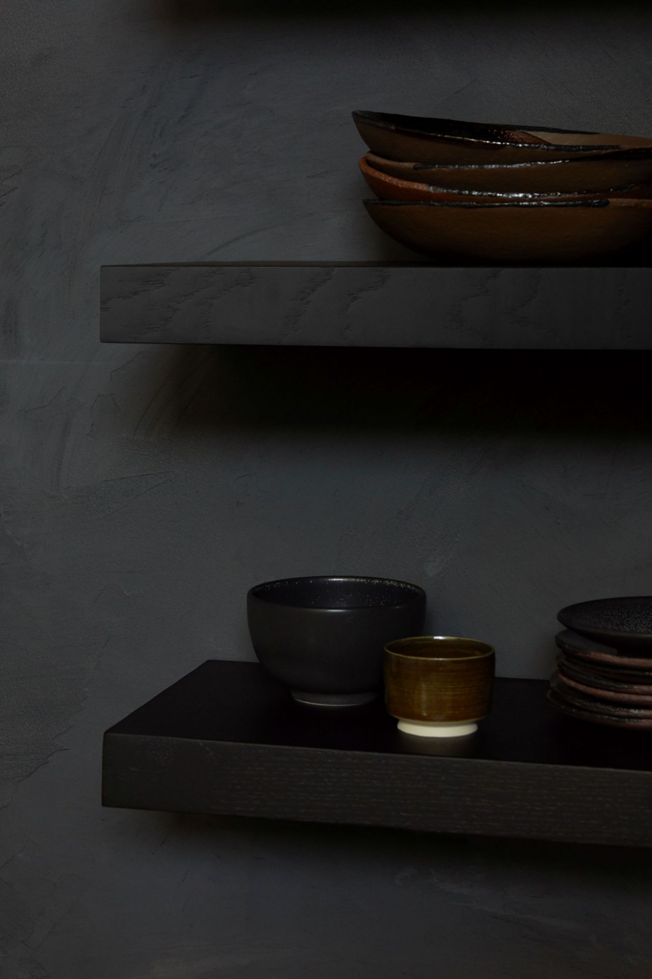 Schwarzes Ethnicraft Wandregal aus Eiche mit Geschirr, Schalen und Tassen auf zwei Ebenen vor dunkler Wand.