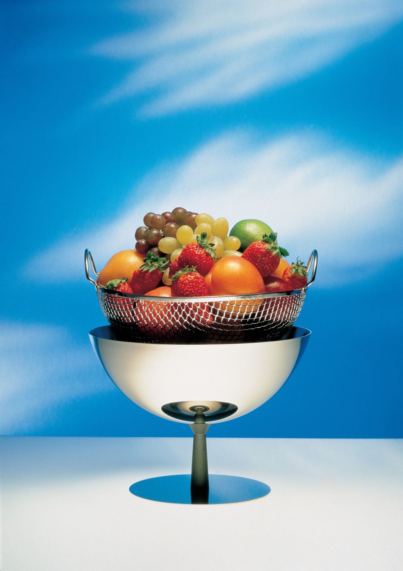 Alessi Obstschale/Sieb AC04 gefüllt mit frischem Obst wie Erdbeeren, Orangen und Trauben. Moderne Küchenschale aus Edelstahl.