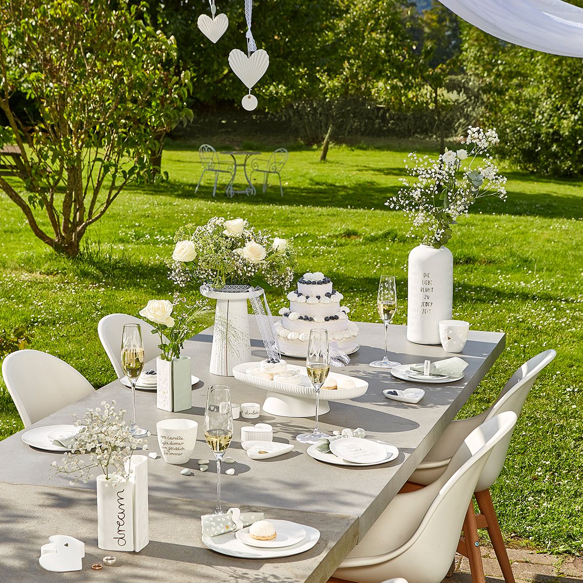 Hochzeitstafel im Garten mit weißem Geschirr, Torte, Champagner und einer plissierten Vase mit Blumen.