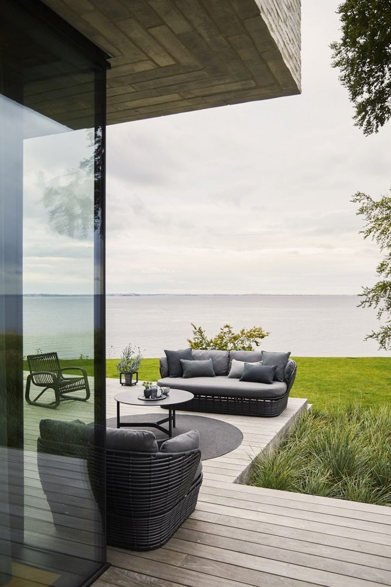 Cane-Line Basket Outdoor Sofa, 2-Sitzer, graues Geflecht mit Kissen auf Holzterrasse am Meer.
