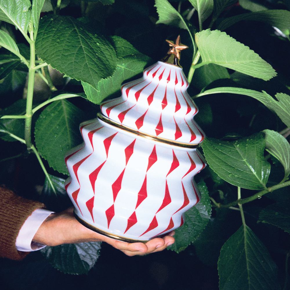 Hand hält Alessi Keksdose NZ02 Delight in Tannenbaumform, weiß mit roten Rauten und goldenem Stern.
