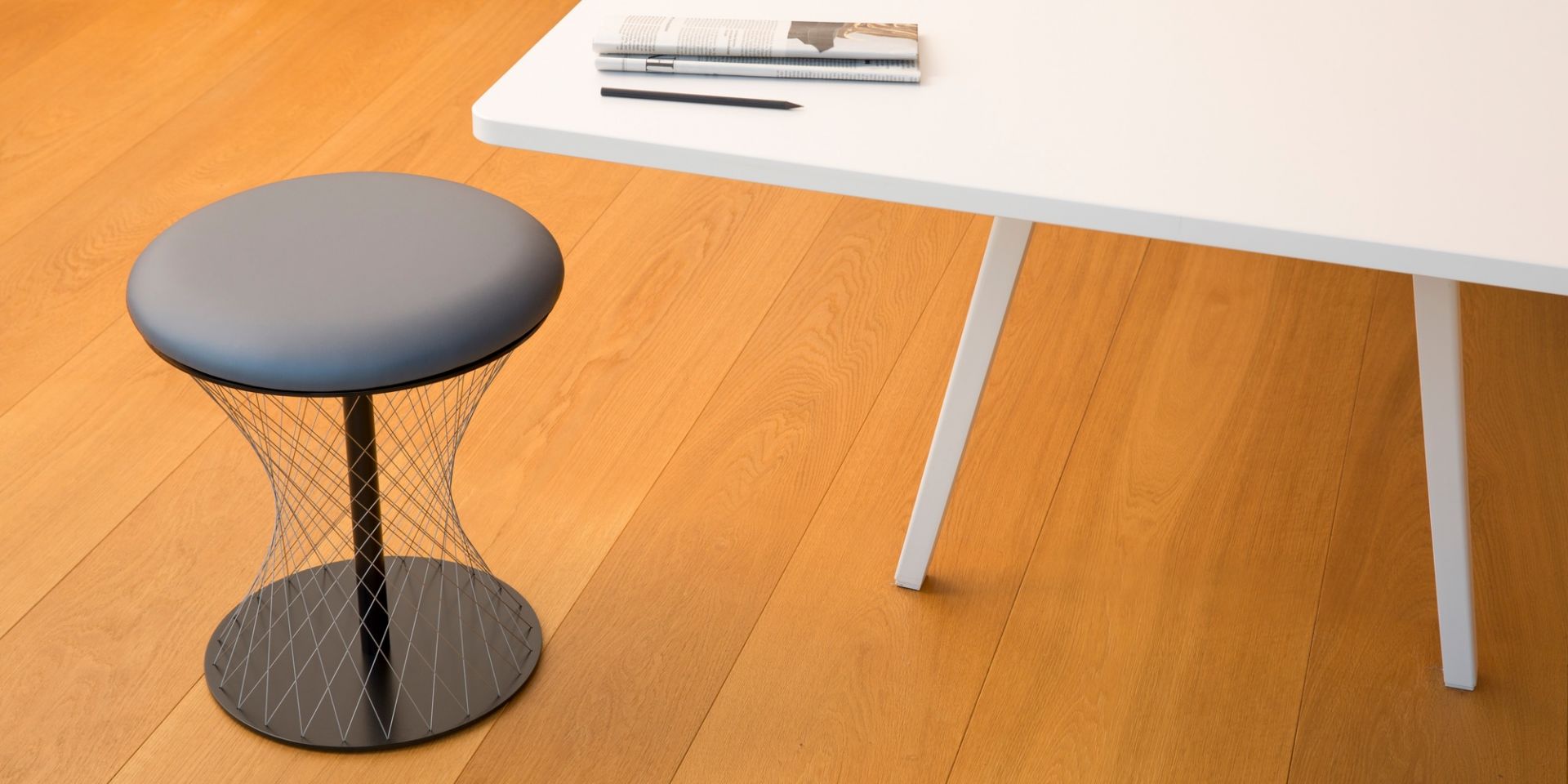 Grauer Network Hocker Favius mit Drahtgestell neben weißem Tisch auf Holzboden.