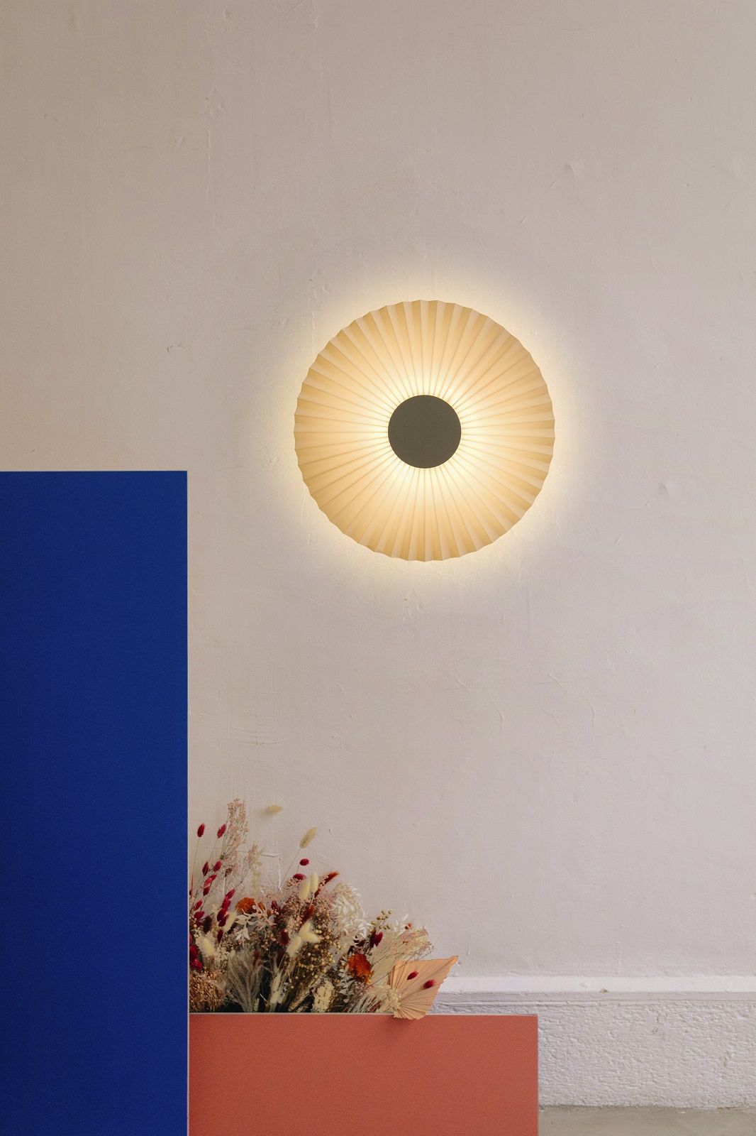 Carmen Wandleuchte von Hartô: Runde, plissierte Designer-Wandlampe in Beige an einer weißen Wand.