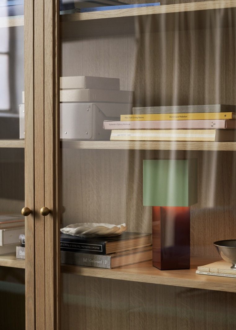 Detailaufnahme des AndTradition Trace Schranks mit Glasfront, Bücher, Lampe und dekorativen Objekten auf Holzregalen.