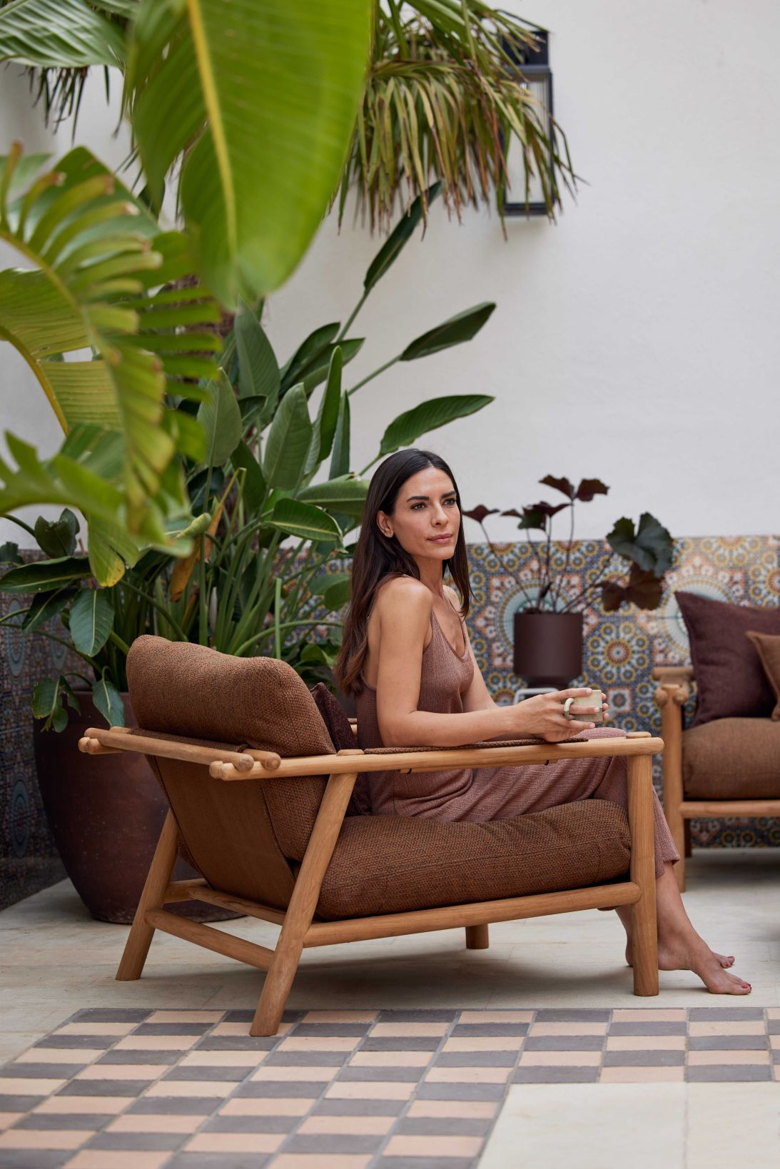 Frau sitzt entspannt im Cane-Line Sticks Outdoor Loungesessel mit Kissen, umgeben von Pflanzen.