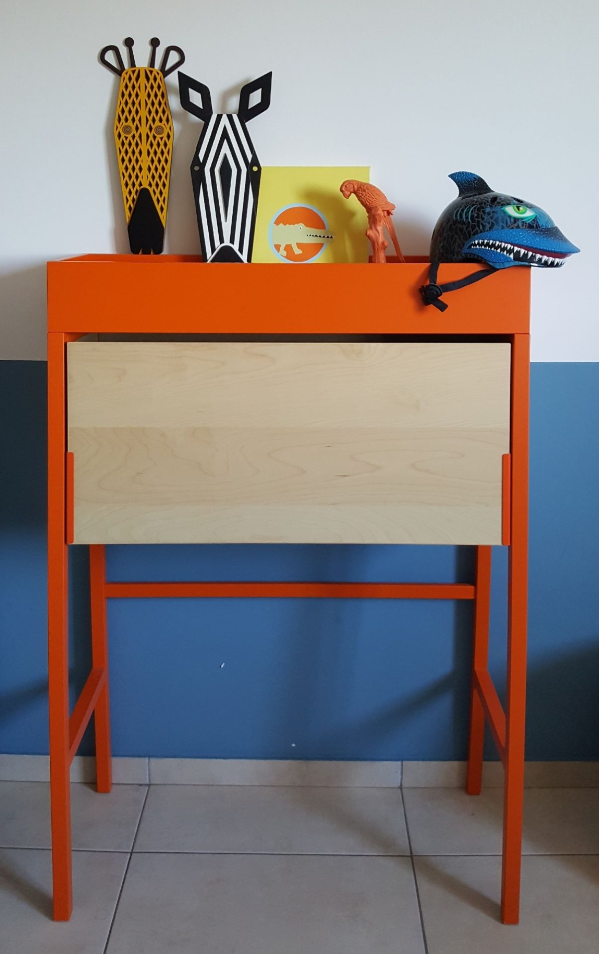 Dekorativer Umasqu Zebra Mini Wandschmuck auf orangefarbenem Tisch, kombiniert mit Giraffenmaske und Hai-Helm als Kinderzimmer-Dekoration.