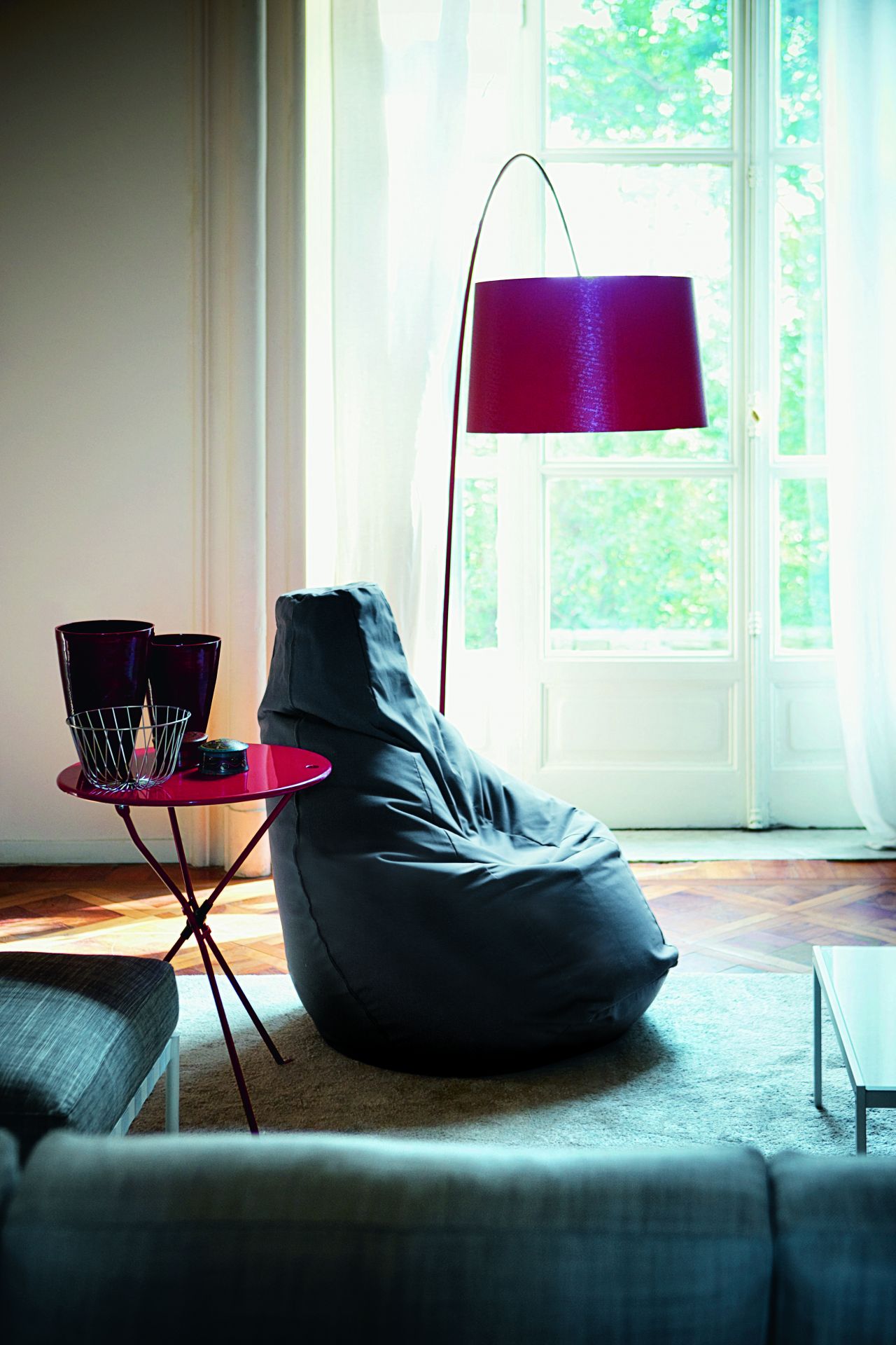 Grauer Sacco Sitzsack von Zanotta neben rotem Beistelltisch und Stehlampe in modernem Wohnzimmer.