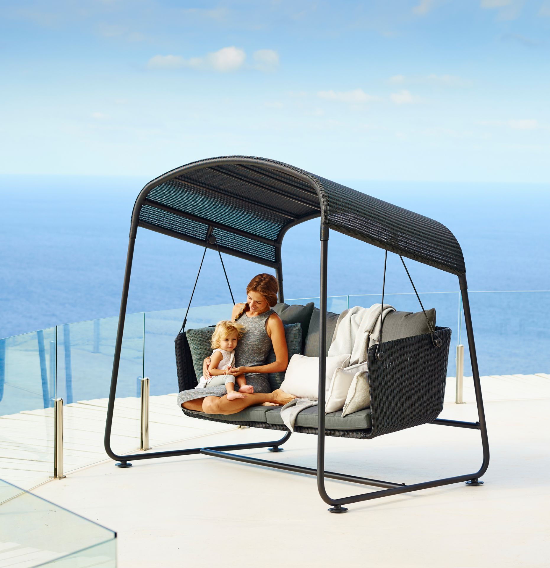 Frau mit Kind auf Cane-Line Cave Gartenschaukel mit Kissen vor Meerblick.